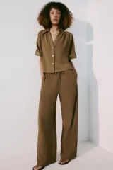 Pantalón de tiro medio color marrón, con cintura elástica y cordón ajustable con abalorios decorativos. Presenta bolsillos laterales y perneras rectas con diseño texturizado.