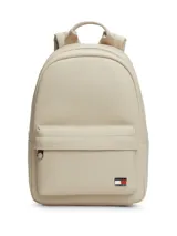 Mochila Tommy Jeans de diseño minimalista en color beige, confeccionada en poliuretano de acabado liso. Cuenta con un compartimento principal con cierre, un bolsillo frontal con cremallera, asa superior y correas ajustables. Incluye el logo metálico de la marca en la parte inferior frontal.
