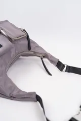 Cartera tipo bandolera de tela técnica color gris, con correa ajustable negra. Presenta dos bolsillos frontales externos: uno con cierre y otro con solapa y hebilla decorativa.