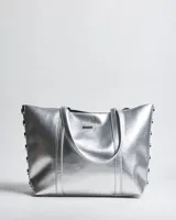 Cartera tipo tote de símil cuero plateado con doble asa de hombro, detalles de tachas en los laterales y placa metálica con logo de la marca en el frente.