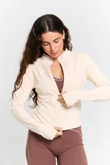 Campera deportiva de ajuste entallado, color crudo, con cuello alto, cierre frontal completo y mangas largas con orificio para el pulgar.