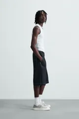 Bermuda de denim rígido color azul oscuro (sin prelavar) con corte amplio (baggy fit). Presenta pliegues frontales, trabillas ajustables en la cintura, bolsillos delanteros y bolsillos traseros de parche, todo resaltado con pespuntes a contraste.