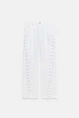 Jeans rectos de tiro medio en color blanco, con cinco bolsillos, trabillas y cierre frontal con cremallera. Presenta detalle de cordones cruzados con ojalillos metálicos a lo largo de los laterales de ambas piernas.
