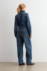 Mono de jean de corte holgado con cierre frontal de botones, cuello camisero y mangas largas. Presenta costuras marcadas y un cinturón de cuero marrón con hebilla metálica en la cintura.