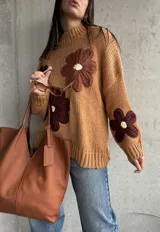 Sweater de punto grueso color camel, con diseño de flores grandes aplicadas en tonos marrones y centro crema. Presenta cuello redondo alto, mangas largas con hombros caídos y terminaciones acanaladas.