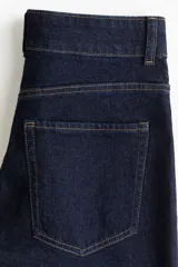 Jean de denim de algodón con leve stretch, color azul oscuro (índigo). Presenta un corte ajustado en la cintura y cadera, ensanchándose en un corte acampanado desde la rodilla hasta el bajo. Es de tiro alto con cierre y botones frontales, y cuenta con solapas decorativas en los bolsillos delanteros y bolsillos parche traseros.
