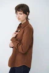 Camisa de estilo casual confeccionada en material tipo gamuza color marrón, con cuello clásico, cierre frontal con botones y costuras a la vista en tono contrastante.