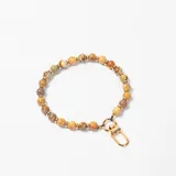 Accesorio decorativo para cartera compuesto por una cadena de cuentas de piedra natural en tonos tierra, intercaladas con pequeñas arandelas doradas, y finalizado con un mosquetón metálico dorado.