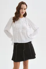 Blusa blanca de viscosa con cuello a la base, mangas largas abullonadas con puños ajustados, detalles de volados, encaje y botones frontales.