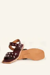 Sandalia baja de cuero vacuno color negro con acabado efecto charol texturizado, decorada con tachas metálicas plateadas en las tiras y la sujección del talón. Cuenta con suela plana de cuero.