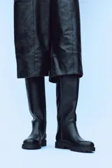 Bota plana de caña alta en color negro, con diseño minimalista y detalle de pespuntes verticales en el cuerpo. Cuenta con suela track, punta redonda y tirador trasero para facilitar el calce.