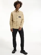 Camisa de manga larga color beige, confeccionada en tejido texturizado de algodón. Presenta cuello camisero, cierre frontal de botones y un bolsillo en el pecho con una insignia cuadrada negra con el logo 'CK' en blanco.