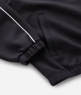 Buzo deportivo de corte boxy, color negro, con cuello alto, cierre frontal completo y mangas largas. Presenta detalles de líneas blancas a lo largo de las mangas y lettering estampado en el pecho.