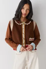 Chaleco de lana color marrón con cuello estilo bebé, cierre frontal con botones grandes y detalle de puntilla en cuello, sisas y bolsillo frontal. Presenta una franja decorativa en color rojo en el ruedo.