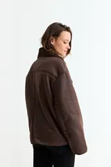 Chaqueta de estilo aviador en color marrón, confeccionada en material simil gamuza con interior y solapas de corderito sintético. Presenta cierre frontal con botones, corte holgado y mangas largas.