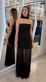 Vestido largo negro strapless con escote palabra de honor, diseño ajustado en el cuerpo y falda larga semitransparente con detalles plisados. Incluye una pieza al cuello tipo choker unida a la parte posterior del vestido.