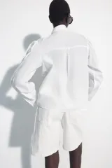 Camisa de algodón color blanco, con cuello subido y detalle de pañuelo incorporado, manga larga con puños anchos y cierre frontal con botones joya dorados.