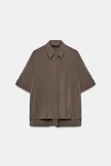 Camisa de corte fluido con cuello solapa y manga corta con dobladillo. Presenta un cierre frontal con botones ocultos bajo una tapeta y un diseño con bajo ligeramente más largo en la parte posterior.