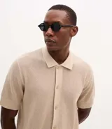 Camisa de punto de manga corta en color beige, con cuello inglés y cierre frontal de botones. Presenta una textura tejida y un corte regular que ofrece un ajuste cómodo y clásico.