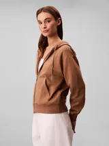 Campera canguro de felpa color marrón, con capucha ajustable con cordones, cierre frontal completo y bolsillos tipo canguro. Presenta el logo retro de Calvin Klein bordado en el pecho.