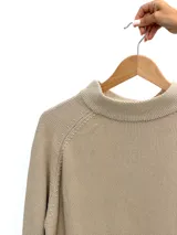 Sweater de punto en color beige, con cuello redondo ligeramente elevado y mangas largas con costura raglán. Presenta un diseño de corte recto con un ruedo asimétrico, siendo más corto en la parte frontal.