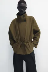 Parka amplia de Zara Woman Collection confeccionada en hilatura con mezcla de algodón, en color verde militar. Presenta cuello alto, manga larga con trabillas y botón, bolsillos laterales ocultos en costura, cintura ajustable con cordones y cierre frontal con botones.