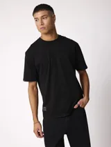 Remera negra de manga corta, cuello redondo y corte holgado, con pequeña etiqueta decorativa en el borde inferior.