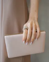 Clutch rígido de acabado satinado en tono champagne, con estructura rectangular y detalles metálicos dorados en los bordes.