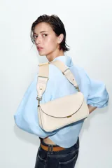 Bolso bandolera alargado de color blanco marfil con solapa redondeada y detalle metálico dorado. Incluye doble asa bandolera ajustable, una de ellas en tejido.