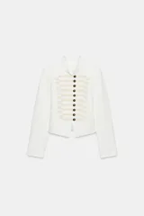 Chaqueta entallada de color blanco con cuello redondo y manga larga. Presenta un diseño de estilo militar con cierre frontal mediante alamares decorativos y botones metálicos.