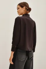 Camisa negra de algodón con cuello alto, cierre frontal de botones y detalles de encaje vertical y en los puños.