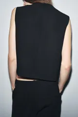 Chaleco negro de cuello redondo y sin mangas, con cierre frontal cruzado con un botón y bajo asimétrico.