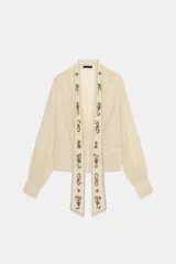 Camisa color crema o marfil, de tejido semitransparente, con escote pico cruzado y mangas largas con puños abotonados. Presenta un detalle de foulard incorporado en el frente, adornado con apliques de abalorios oscuros en forma de motivos florales y geométricos.