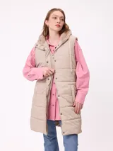 Chaleco largo acolchado color beige, con capucha forrada en sherpa, cierre frontal con cremallera y botones a presión, y bolsillos laterales.