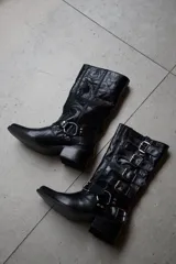 Bota estilo biker de caña alta, confeccionada en cuero vacuno color rojo cherry. Presenta un diseño con múltiples tiras horizontales con hebillas metálicas, punta redondeada y taco cuadrado bajo de madera.