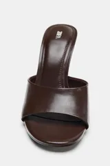 Zapato tipo mule de piel color marrón, con tacón de cuña asimétrica de madera y acabado en punta redonda.