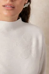 Buzo de punto color visón con cuello alto, manga larga y diseño holgado. Presenta un bordado floral en relieve al tono en la parte frontal.