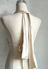 Top color crema o amarillo pálido, estilo halter con cuello atado y escote pronunciado en V con drapeado frontal. El cuello está adornado con una flor de orquídea en tonos amarillo, naranja y rosa.