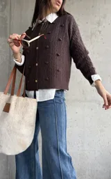 Cardigan de punto color chocolate, con diseño de trenzas y pompones decorativos. Presenta cuello redondo, cierre frontal con botones metálicos dorados y mangas largas.