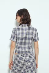 Camisa de manga corta con cuello camisero y botones frontales. Presenta un estampado de cuadros escoceses en tonos marrón oscuro y blanco/beige, con un calce entallado.