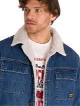 Chaqueta de denim azul con forro interior de sherpa color crema. Presenta cierre frontal con botones metálicos, bolsillos superiores con solapa y bolsillos laterales tipo welt. Incluye etiqueta con logo de la marca en el bolsillo del pecho.