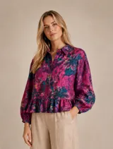 Camisa de lino con estampado floral en tonos fucsia, violeta y azul. Presenta cuello clásico, cierre frontal con botones, mangas largas abullonadas con puños elásticos y un detalle de volado en el ruedo.