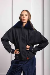 Campera oversize de jean color negro con cuello de corderito azul, cierre frontal con cremallera y solapa con botones a presión. Presenta mangas amplias y un pequeño bordado rojo en el puño.