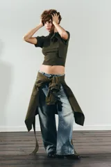 Jersey de punto de manga corta y cuello redondo, en color verde oliva. Presenta un detalle de lazo grande en el frente que le otorga un toque distintivo y femenino.