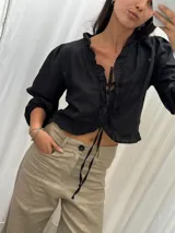 Blusa negra de lino con acabado brillante, de manga larga abullonada, escote con volados y cierre frontal con tiras para anudar.