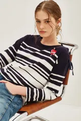Remera de manga larga a rayas horizontales en azul marino y blanco, con cuello redondo y detalle de parche bordado en forma de corazón con flecha en el pecho. Presenta lazos decorativos en los hombros.