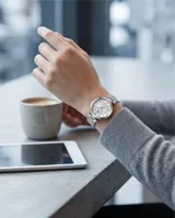 Reloj de pulsera analógico con caja redonda de metal plateado y malla de acero tipo milanesa. Presenta una esfera minimalista con números romanos y manecillas finas.