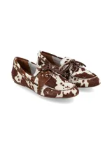 Mocasín de mujer con estampado de vaca (pelo marrón y blanco), estilo náutico o driver, con cordones marrones decorativos en el empeine y ojales metálicos dorados.