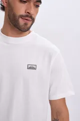 Remera blanca de manga corta con cuello redondo y una pequeña etiqueta estampada en el pecho con diseño de montaña y texto.