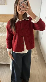 Cardigan de punto con textura trenzada en color bordó, con cuello redondo y cierre frontal mediante botones tipo alamar. Presenta mangas largas con puños acanalados y un corte clásico.